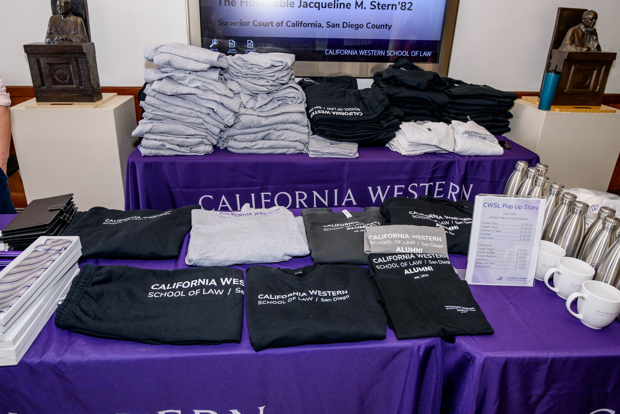Tables with purple tablecloths featuring the CWSL logo are filled with California Western School of Law merchandise. From top left to bottom right, there are: piles of gray, black and white clothes, gray and purple scarves, black padfolios, black sweat pants, black sweatshirts and hoodies, gray hoodies, gray t shirts, a pricing sign, white mugs and stainless steel waterbottles. 